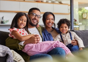 Família sorridente sentada no sofá de casa, transmitindo alegria, união e bem-estar.