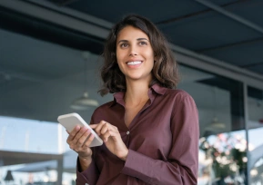 Mulher sorridente usando um celular enquanto está ao ar livre, transmitindo leveza e modernidade.
