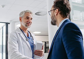 Um médico de jaleco branco cumprimenta um homem de terno azul em um ambiente profissional, sugerindo uma reunião ou conversa formal em uma clínica ou hospital.